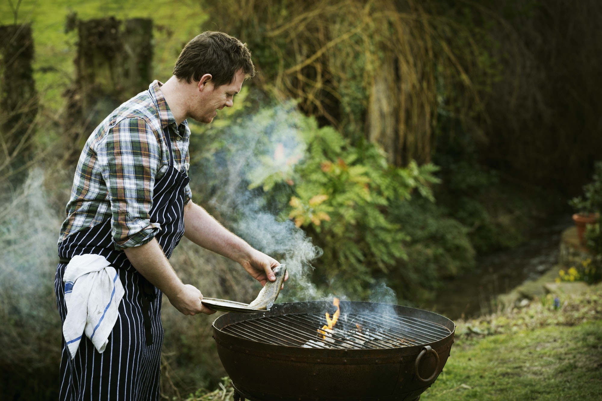 Chef standing in a garden, grilling fish on a barbecue.